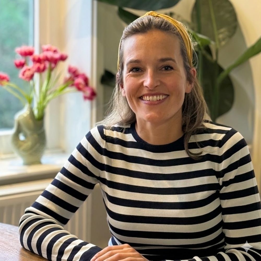 Portretfoto van Chris, expert in cultuurtransformatie en neurodiversiteit, zittend aan een houten tafel en lachend in een kamer met planten en roze tulpen. Ze draagt een geelgebreide hoofdband en een zwart-wit gestreepte trui. (Zichtbaar op de achtergrond zijn ook een radiator en een raam).